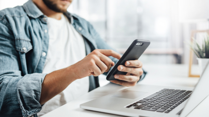 man holding a mobile phone while sitting in front of a laptop