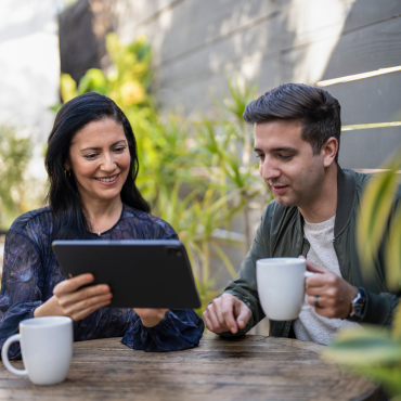 A Hispanic couple sips coffee at their porch table, reading some materials on a tablet computer.